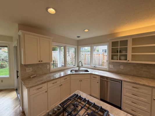 Modern kitchen in Mills with white cabinets, marble countertops, stainless steel appliances, and an island stove. Large windows look out on fenced backyard. Available April 2025. For painting needs in Portland, contact Pearl Painters—experienced painters for interior spaces.
