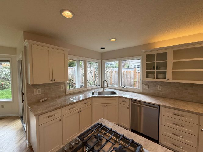 Modern kitchen in Mills with white cabinets, marble countertops, stainless steel appliances, and an island stove. Large windows look out on fenced backyard. Available April 2025. For painting needs in Portland, contact Pearl Painters—experienced painters for interior spaces.