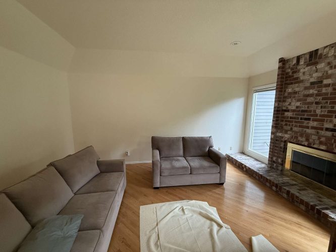 A painter from Pearl Painters in Portland is working in a cozy living room with two gray sofas, a brick fireplace, and a window. A sheet is partially spread on the wooden floor. The plain walls reflect the simple interior painting trends of April 2025.