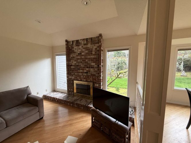 A living room in Mills features a brick fireplace, two large windows, a gray sofa, and hardwood floors. Pearl Painters, Portland painting professionals, served this interior.