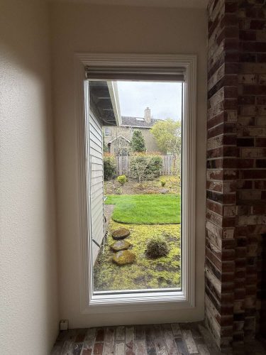 A painting by Pearl Painters, Portland, shows a green backyard with stepping stones, a wooden fence, and neighboring houses as seen through a rectangular window. Part of an indoor brick wall is also visible. Inspired by Mills, April 2025.