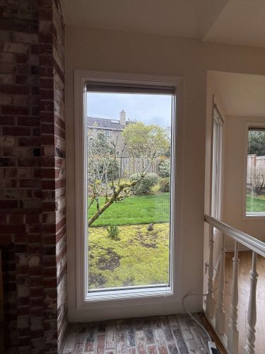 Painting by Mills, April 2025. Depicts a large rectangular window in a white interior wall beside a brick wall, overlooking a green backyard with trees and shrubs. Portland. Pearl Painters.