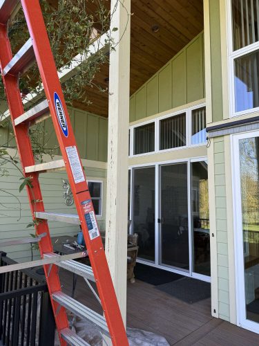 A red ladder leans against the green exterior of a house with large glass doors and vertical siding. A pillar appears prepped for repair, indicating upcoming painting work by Pearl Painters in Portland, likely part of McCaffery’s spring 2025 projects.
