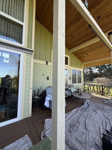 A painter from Pearl Painters in Portland works on a covered exterior patio with light green siding, wooden ceiling, and large windows. Protective tarps cover the floor during this April 2025 painting update.