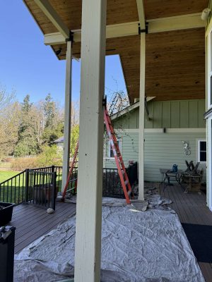 In April 2025, a painter from Pearl Painters in Portland set up a red ladder against the green exterior of a house under a covered porch with exposed beams and tarps on the deck, preparing for painting.