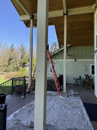 In April 2025, a painter from Pearl Painters in Portland set up a red ladder against the green exterior of a house under a covered porch with exposed beams and tarps on the deck, preparing for painting.