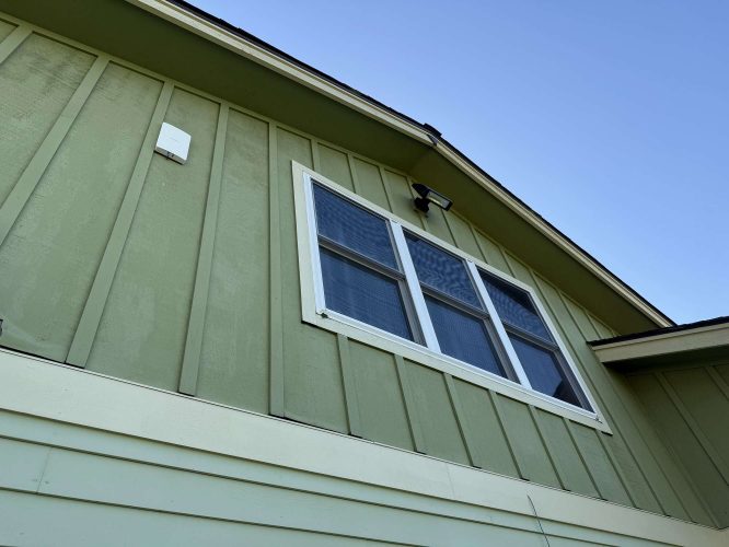 A painter photographed the exterior of a green house with vertical siding, a white-framed window, and a security camera in Portland for Pearl Painters in April 2025. A small white device is mounted on the wall under a clear blue sky.
