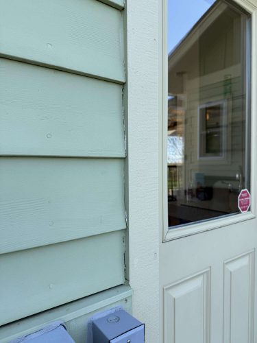 Close-up of a light green exterior wall with horizontal siding painted by Pearl Painters in Portland, beside a cream-colored door with a window reflecting a house and yard, April 2025.