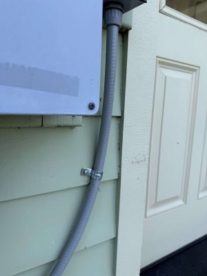 A gray electrical conduit is attached to a green exterior wall near a white door and utility box—exterior painting work by McCaffery, painter, in Portland, April 2025. Pearl Painters.