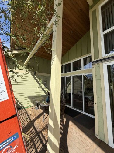 A painter from Pearl Painters in Portland photographed a green house with a sloped wooden ceiling, large windows, and a sliding glass door. A red ladder is on the left side of the covered porch. Photo taken April 2025.