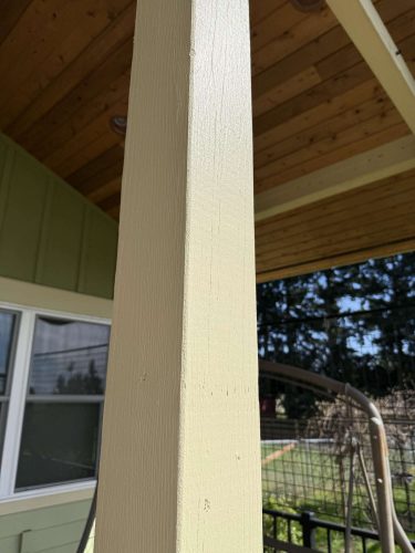 Close-up of a beige-painted wooden porch post in front of a green house wall, with window and wooden ceiling. Painting by Pearl Painters, Portland. Photo by McCaffery, April 2025.
