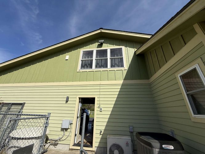 Photo of a light green house exterior with two upper windows, HVAC units, and a fence in Portland, taken by painter McCaffery in April 2025. Pearl Painters project.