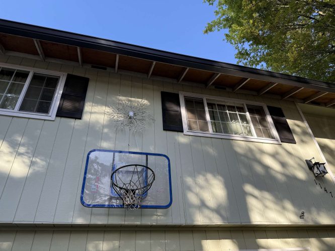 A Wehage basketball hoop is mounted below two windows on a house exterior in Portland. There is damage above the hoop. Trees are visible top right. Photo taken April 2025. Pearl Painters, a local painting company, can repair and repaint this area—call for a painter quote.