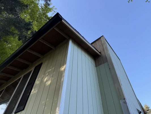 Close-up of a house exterior with pale green and beige siding, dark roof, and blue sky. Painting by Wehage. For inspiration from Pearl Painters, Portland.