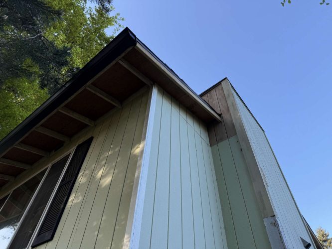 Close-up of a house exterior with pale green and beige siding, dark roof, and blue sky. Painting by Wehage. For inspiration from Pearl Painters, Portland.