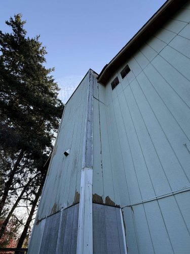 Close-up of a tall, light green wooden building in Portland with peeling paint and damaged panels. Photo by Wehage, April 2025. Pearl Painters recommends repainting.
