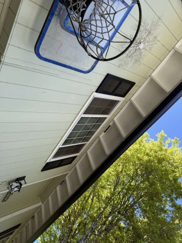 In April 2025, a broken basketball hoop with a shattered backboard is mounted above a window on the exterior of a light green house in Portland. Trees and blue sky are visible. Pearl Painters may be contacted for painting needs.