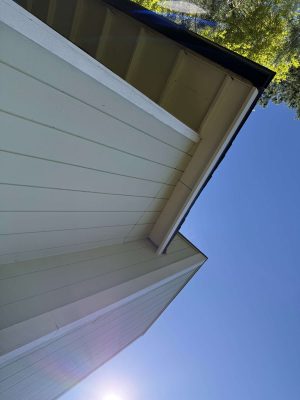 A close-up of a house’s exterior corner, eaves, and roof displays Wehage painting craftsmanship in Portland under a clear blue April 2025 sky, with tree branches in the top right.