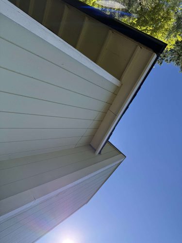 A close-up of a house’s exterior corner, eaves, and roof displays Wehage painting craftsmanship in Portland under a clear blue April 2025 sky, with tree branches in the top right.