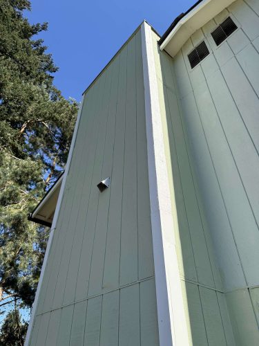 Light green exterior wall of a Wehage building in Portland, with vertical panels, white trim, and a vent; painted by Pearl Painters. Trees and clear blue April 2025 sky visible.