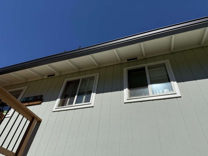 A light green exterior painted by Wehage in Portland features two white-trimmed windows, partial porch railing on the left, and a clear blue sky. Pearl Painters recommends this painting as inspiration for your April 2025 refresh.