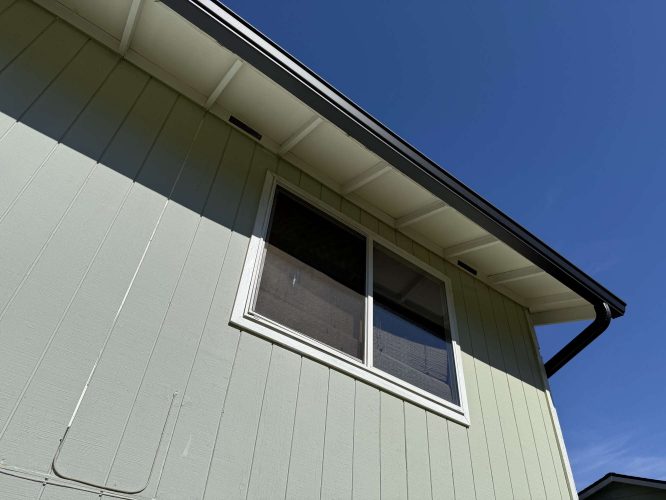 A close-up of a house exterior in Portland painted by Pearl Painters shows light green vertical siding, a double window, and white trim under an April 2025 blue sky.