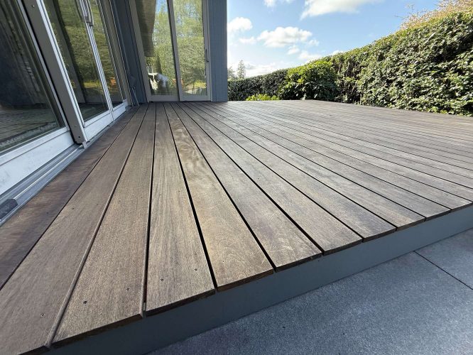 A painter from Pearl Painters in Portland worked on a wooden deck extending from a glass patio door. Surrounded by green hedges under a partly cloudy sky, the painting will be completed by April 2025.