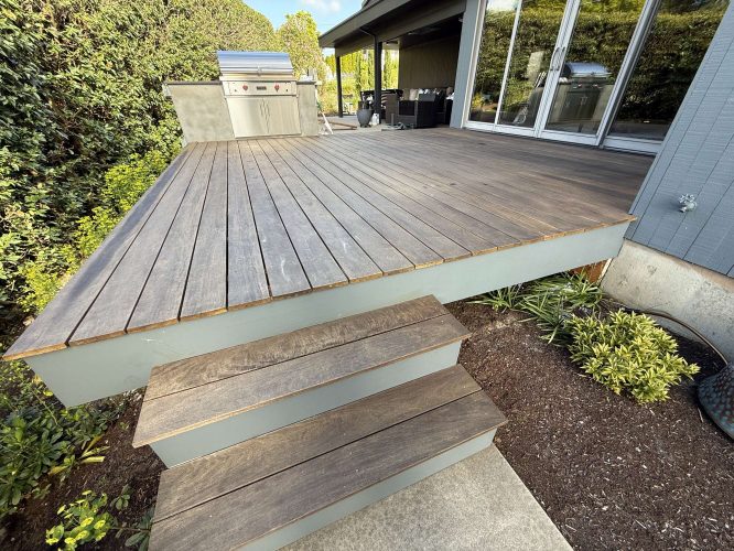 A wooden deck with two steps leads to a grill and outdoor kitchen area next to a house with sliding glass doors and plants. Pearl Painters, Portland painters, can refresh this outdoor space with professional painting before spring gatherings in April 2025.