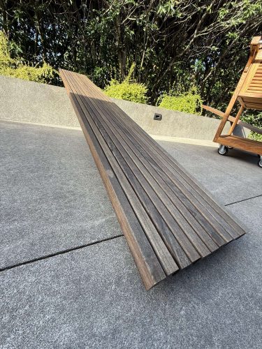 This painting by Pearl Painters of Portland shows a long, narrow wooden bench with a slatted surface on a concrete patio in the Godbey exterior. Lush greenery and a wooden cart are visible in the background. Painted in April 2025.