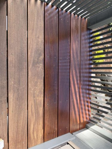 Photo of four vertical wooden boards from the Godbey Exterior project in Portland, April 2025. The two right boards are freshly stained darker; the two left boards are lighter and unstained. Sunlight creates slatted shadows. Work by Pearl Painters, Portland painters.