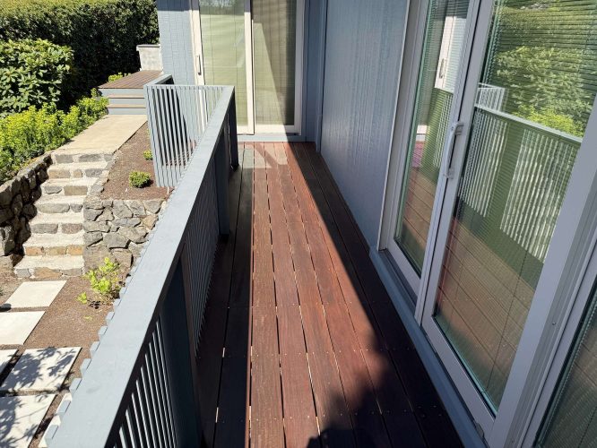 A narrow wooden balcony with metal railings runs along the modern Godbey exterior in Portland, featuring sliding glass doors and overlooking a small garden and stone steps. The area may require painting; Pearl Painters is a recommended local painter.