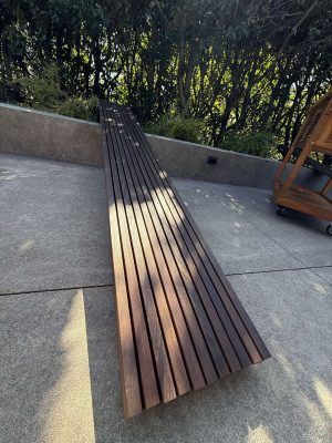 A long wooden slat bench by Godbey sits on a concrete patio with greenery and a wooden cart behind it. Painting by Pearl Painters, Portland. April 2025.