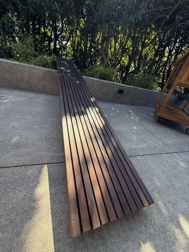A long wooden slat bench by Godbey sits on a concrete patio with greenery and a wooden cart behind it. Painting by Pearl Painters, Portland. April 2025.