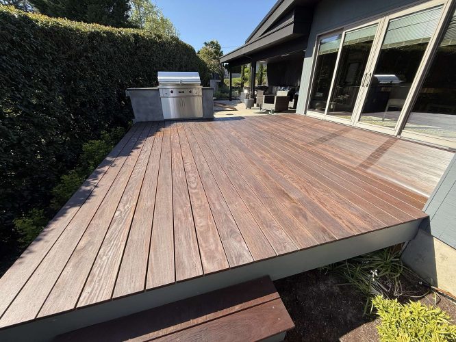 A wooden patio deck in Portland next to a house with large glass doors features a built-in stainless steel grill and lush greenery. Pearl Painters can paint this outdoor area, inspired by Godbey designs, for use in April 2025.