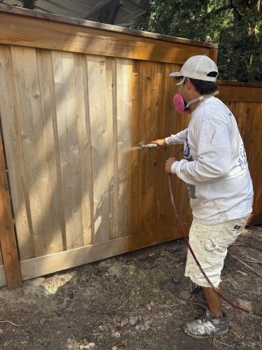 Painter Smith with protective gear stains a wooden fence using a spray gun for Pearl Painters, Portland, July 2025.
