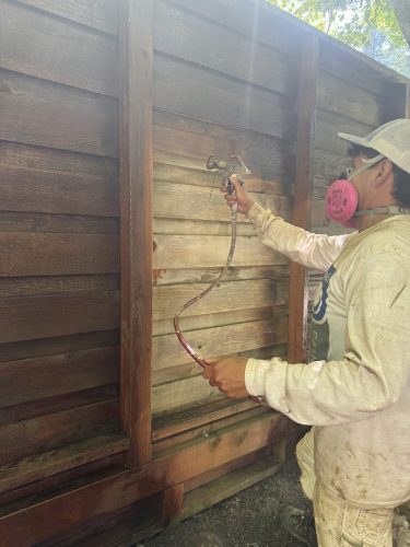 In July 2025, painter Harvey from Pearl Painters sprays exterior paint on a wooden fence in Portland while wearing safety gear.