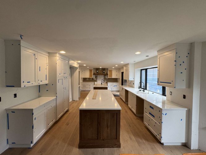 A modern kitchen in Evans, not Portland, is under construction with white cabinets, blue painter’s tape, a wood island, light wood floors, and a large window. Pearl Painters will complete painting by April 2025.
