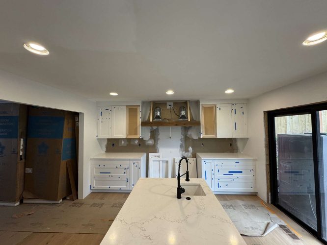 Kitchen renovation in progress with white cabinets partially installed by Evans, blue painter’s tape on cabinet fronts, a center island with sink, and unfinished walls near the stove. Painting work by Pearl Painters in Portland.