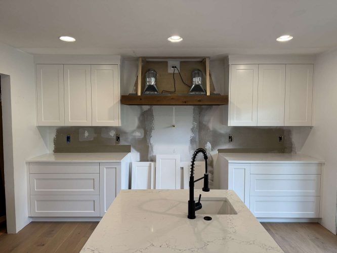 White Evans kitchen in Portland is under renovation: cabinet doors are removed, the wall behind the range hood is exposed, and a central island with a black faucet and sink shows off the progress by Pearl Painters.