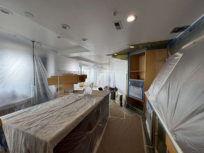 In April 2025, a kitchen in Portland is being renovated. Cabinets, appliances, and surfaces are covered with plastic sheets and tape. Painter tools and materials from Pearl Painters are scattered throughout the room.
