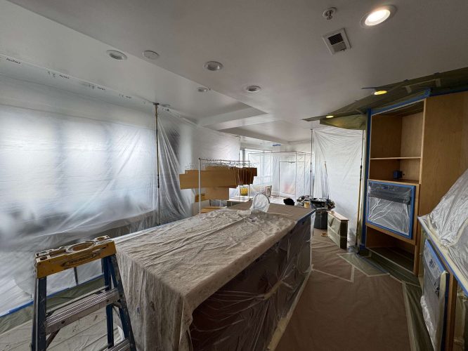 A kitchen in Portland is under renovation, with cabinets and appliances covered for painting. Tools and materials are scattered, and a ladder is set up for Pearl Painters. Work to be completed by April 2025.