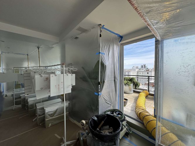 In April 2025, Pearl Painters worked on a painting project in Portland for Isadore. The room under renovation featured plastic sheeting, drying racks with paper, cabinets not yet installed, a trash bin, and a yellow ventilation hose leading to a balcony overlooking the city.