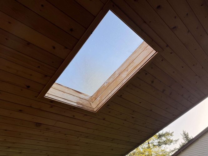 A painter from Pearl Painters in Hamilton painted a rectangular skylight set in a wooden ceiling, showing the sky and tree tops, April 2025.