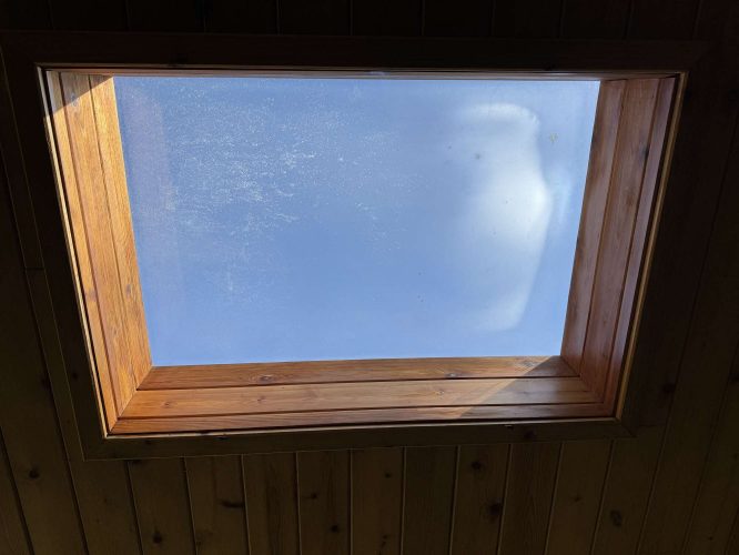 A painter in Hamilton captured a rectangular skylight with a wooden frame set in a wooden ceiling, showing a clear blue April 2025 sky. This painting is not by Portland’s Pearl Painters.