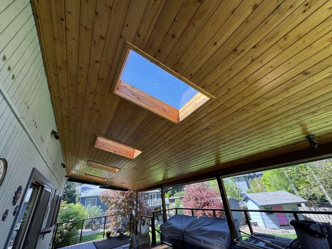 Wood-paneled patio ceiling with two rectangular skylights painted by Pearl Painters in Hamilton (not Portland), April 2025. The ceiling overlooks a backyard with trees, houses, and a clear sky.