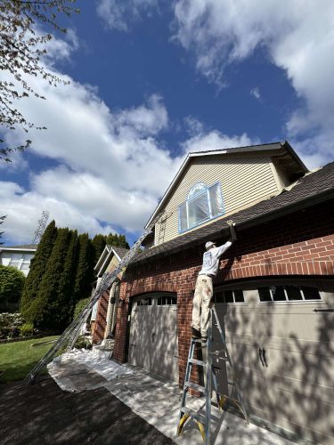 In April 2025, painter Scott of Pearl Painters paints exterior trim near the roof of a two-car garage attached to a brick house in Portland.