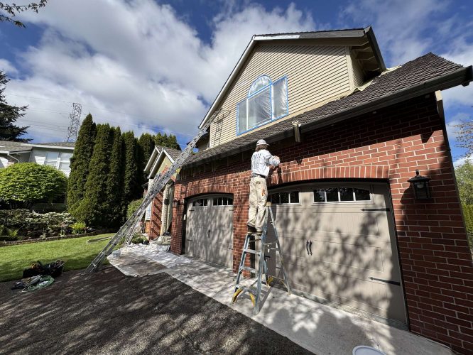 Painter Scott stands on a ladder painting exterior trim above garage doors of a brick and siding house in Portland for Pearl Painters in April 2025.