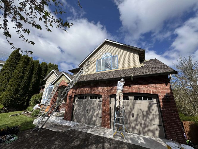 Painter Scott stands on a ladder painting exterior siding above the garage of a two-story brick house in April 2025. This work is done by Pearl Painters in Portland under partly cloudy skies.