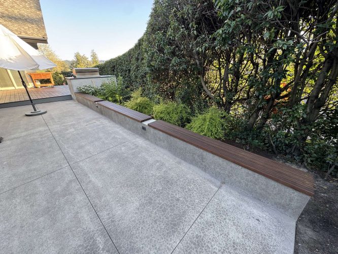 Modern exterior concrete patio with built-in bench seating and wood slats, surrounded by green hedges and shrubs, next to a Godbey-designed house in Portland. Pearl Painters can paint or maintain this area for a polished outdoor look. Ideal for enjoying April 2025 outdoors.