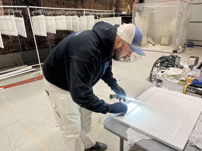 Painter Evans, wearing a hoodie, cap, and gloves, works on cabinets under a flashlight in his Portland workshop. Newly painted doors from April 2025 hang drying in the background. Pearl Painters.